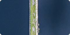 Aerial view of a narrow strip of land with grass and rocks, bordered by water on both sides, with a few people walking along the path.