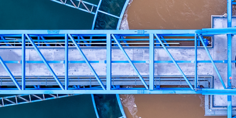 Aerial view of a blue metal bridge structure over water, with one side greenish and the other brown, possibly indicating different water sources or treatment stages.